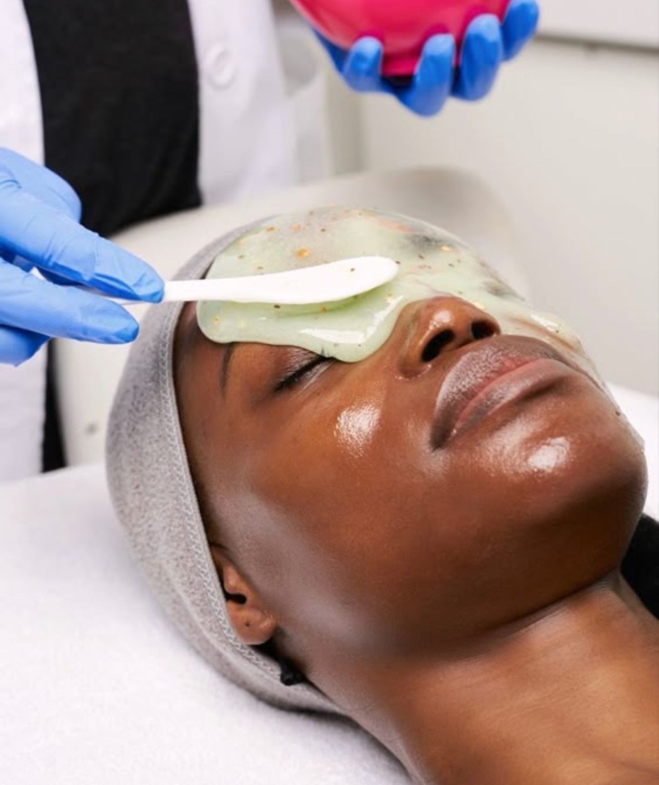 Person receiving a facial treatment with a gel mask applied by a cosmetologist wearing blue gloves.